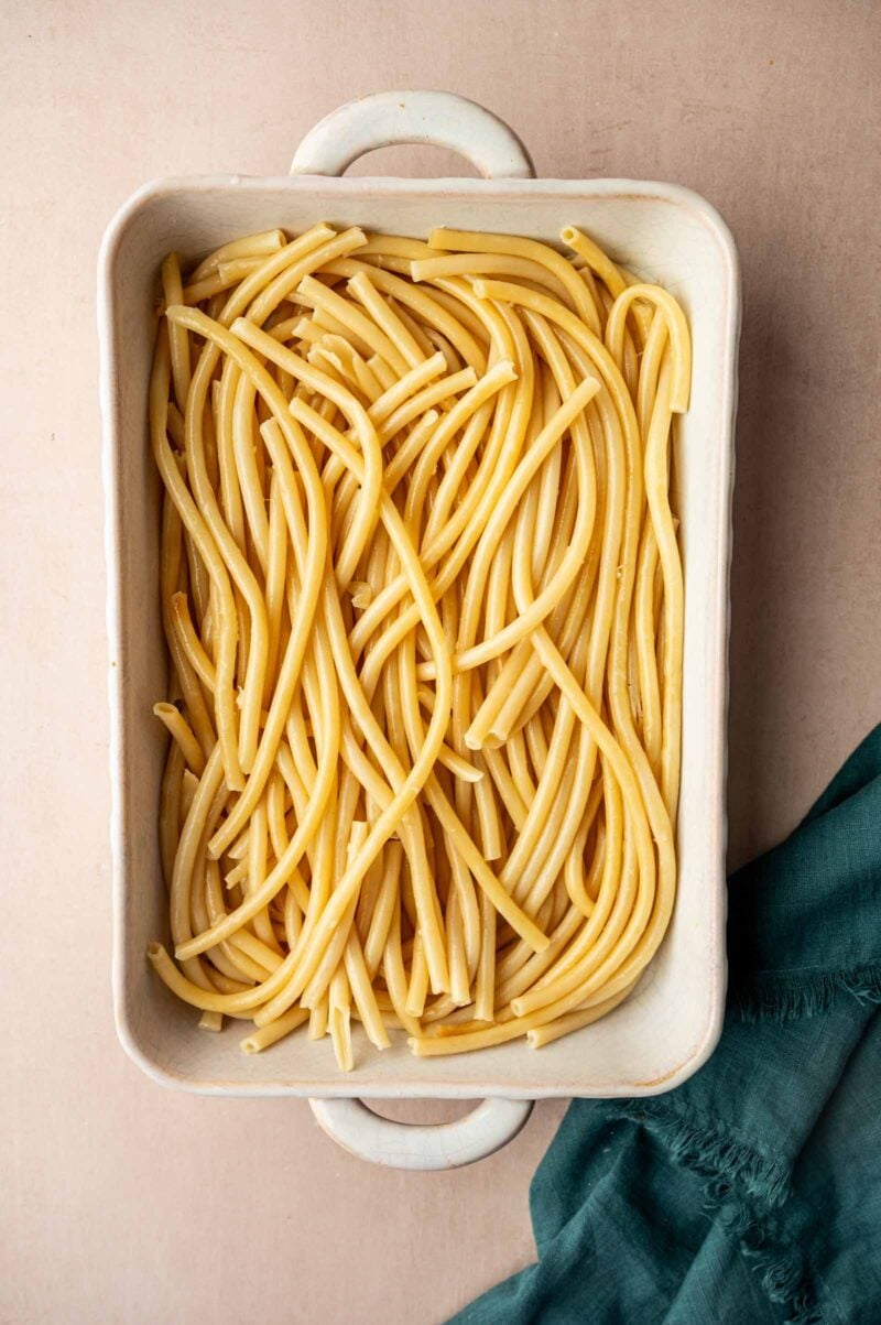 A white rectangular baking dish filled with cooked bucatini pasta sits on a light surface next to a dark green cloth.