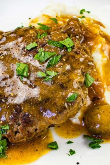 A plate of Salisbury steak with brown gravy, mashed potatoes, whole mushrooms, and chopped parsley garnish.