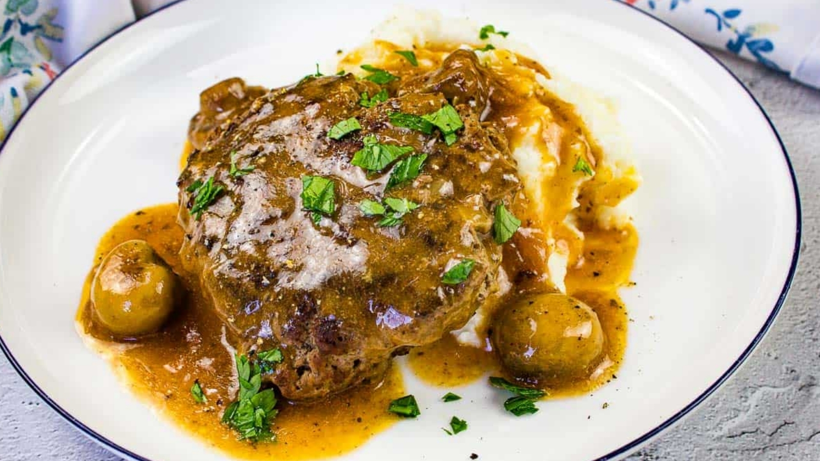 A plate of Salisbury steak with brown gravy, mashed potatoes, whole mushrooms, and chopped parsley garnish.