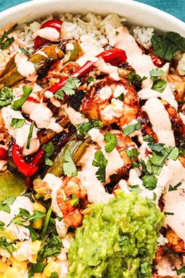 A bowl of rice topped with grilled chicken, vegetables, corn, guacamole, lime wedges, chopped cilantro, and a creamy sauce, placed on a green napkin.