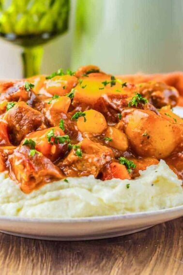 A plate of mashed potatoes topped with chunks of meat, carrots, and mushrooms in a brown sauce.