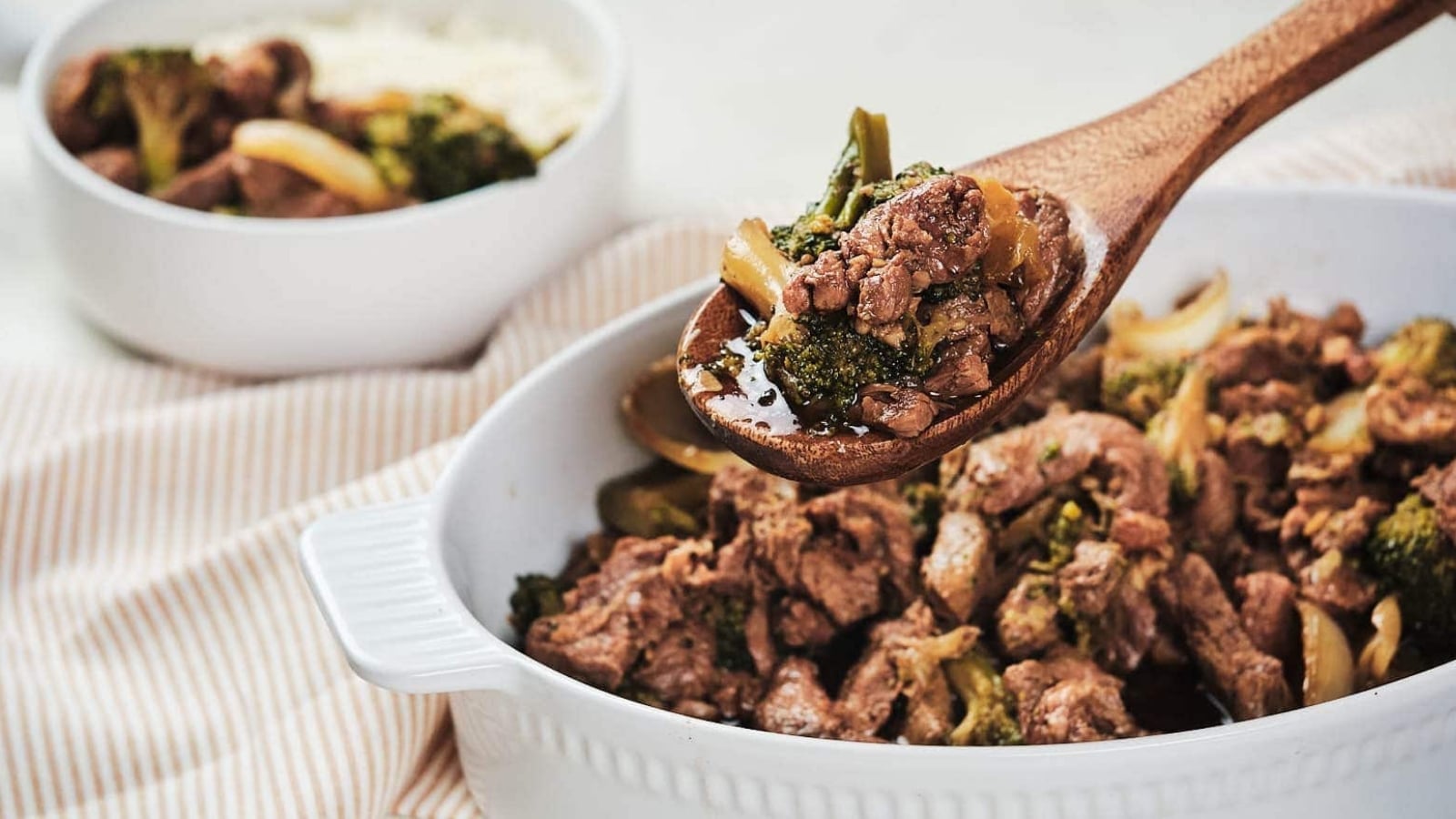 A wooden spoon holds a portion of beef and broccoli stir-fry above a white dish filled with the same food; a similar dish is visible in the background.