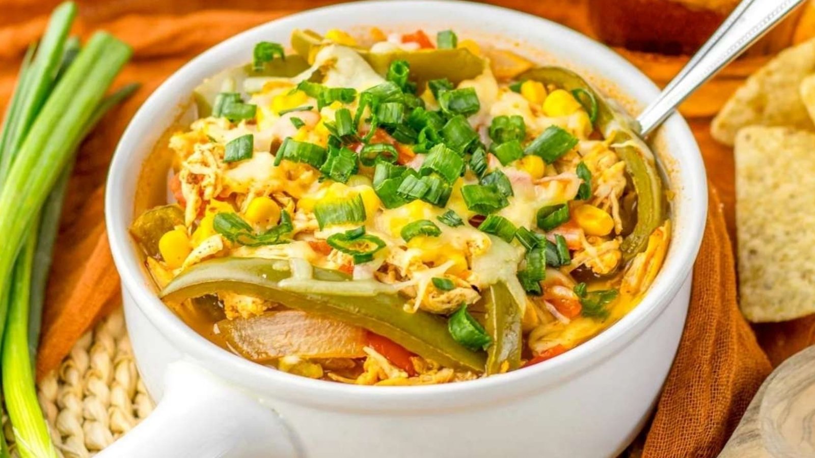 A white bowl filled with shredded chicken, corn, sliced green peppers, melted cheese, and chopped green onions, with a spoon inside.