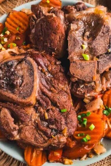 A plate of cooked meat with bone, served on top of sliced carrots, garnished with chopped green onions. Utensils and seasoning bowls are placed nearby on a burlap surface.