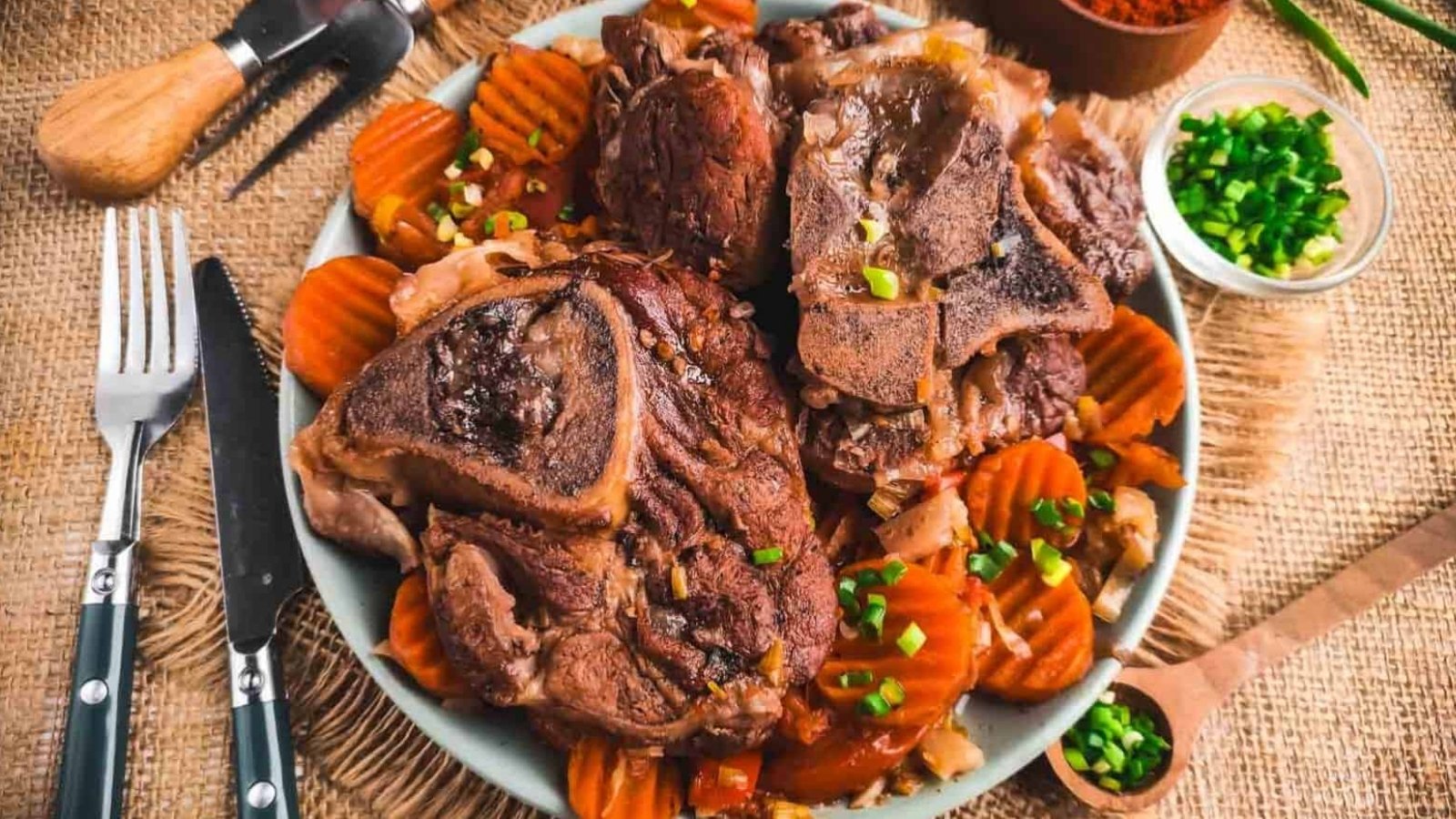 A plate of cooked lamb chops sits on a bed of sliced, cooked carrots. The dish is garnished with chopped green onions. Utensils, a wooden spoon with spices, and a small bowl of more green onions are nearby on a burlap cloth surface.