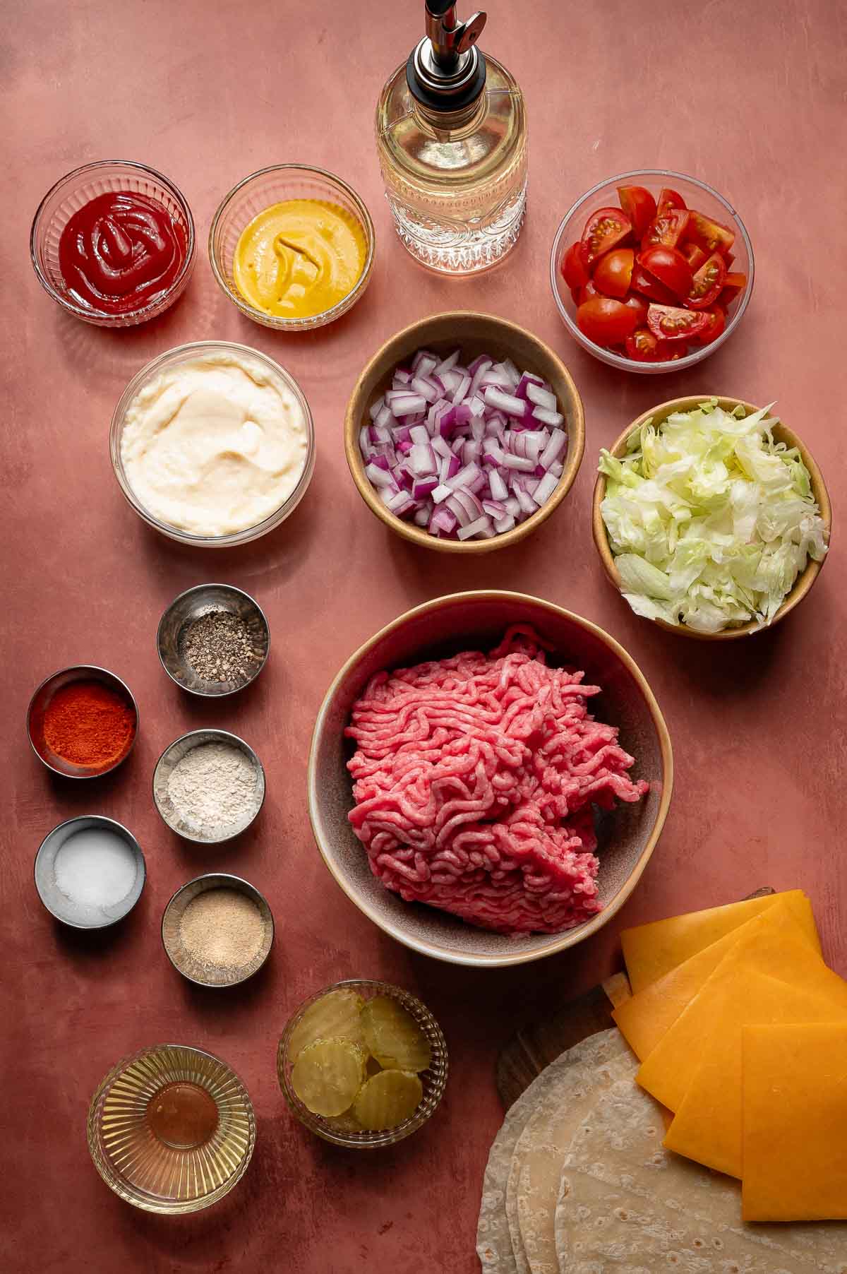 Ingredients for smash burger tacos with special sauce on a flat lay; A bowl of raw ground beef surrounded by sliced cheddar, lettuce, tomatoes, diced onions, pickles, condiments, spices, and tortillas.