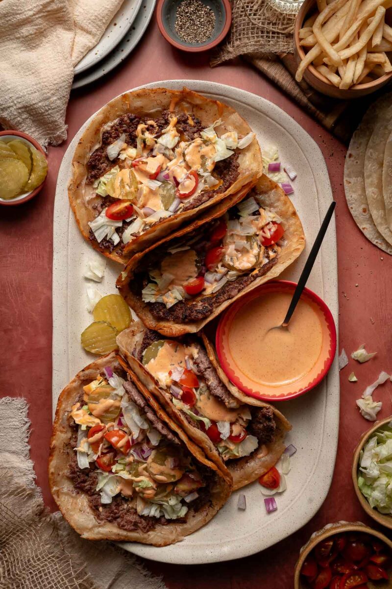 Three smash burger tacos filled with smashed beef, lettuce, tomato, onions, and special sauce are served on a white platter with pickles, a bowl of sauce, fries, and additional toppings on the side.