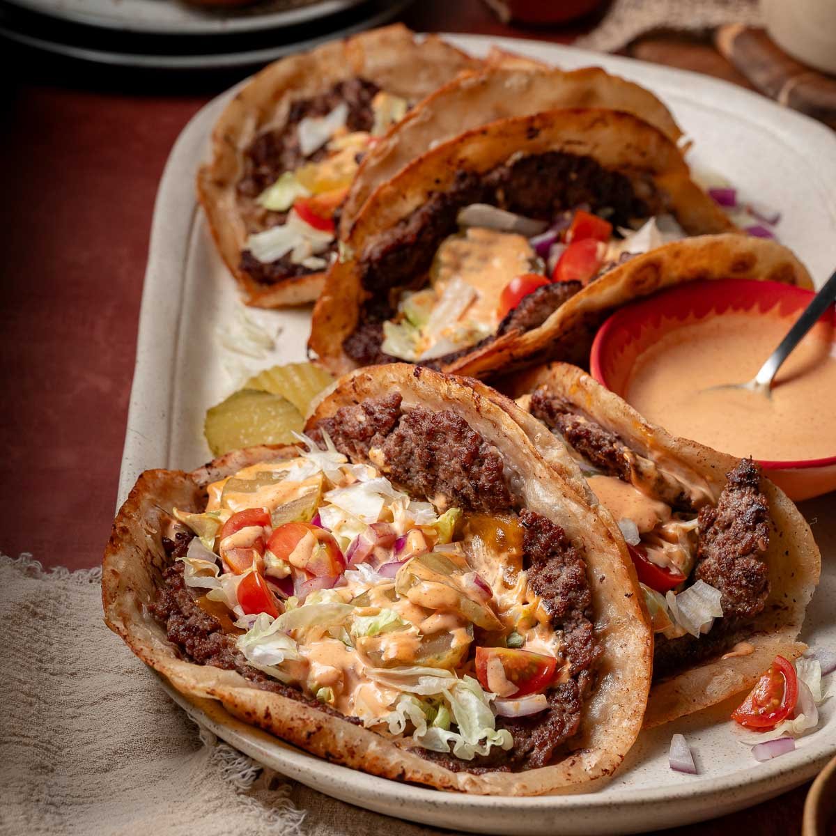Three cheeseburger tacos with ground beef, lettuce, tomato, onions, cheese, and sauce are served on a rectangular plate with a side of special sauce and pickle slices.