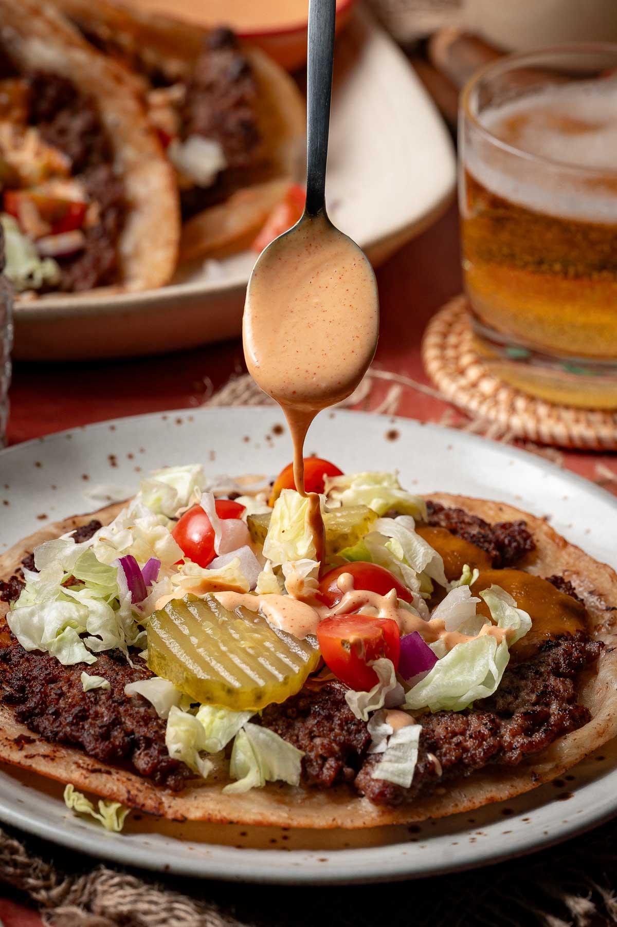 A spoon drizzles special sauce over a smash burger taco topped with ground beef, lettuce, pickles, tomatoes, and onions on a plate, with a drink in the background.