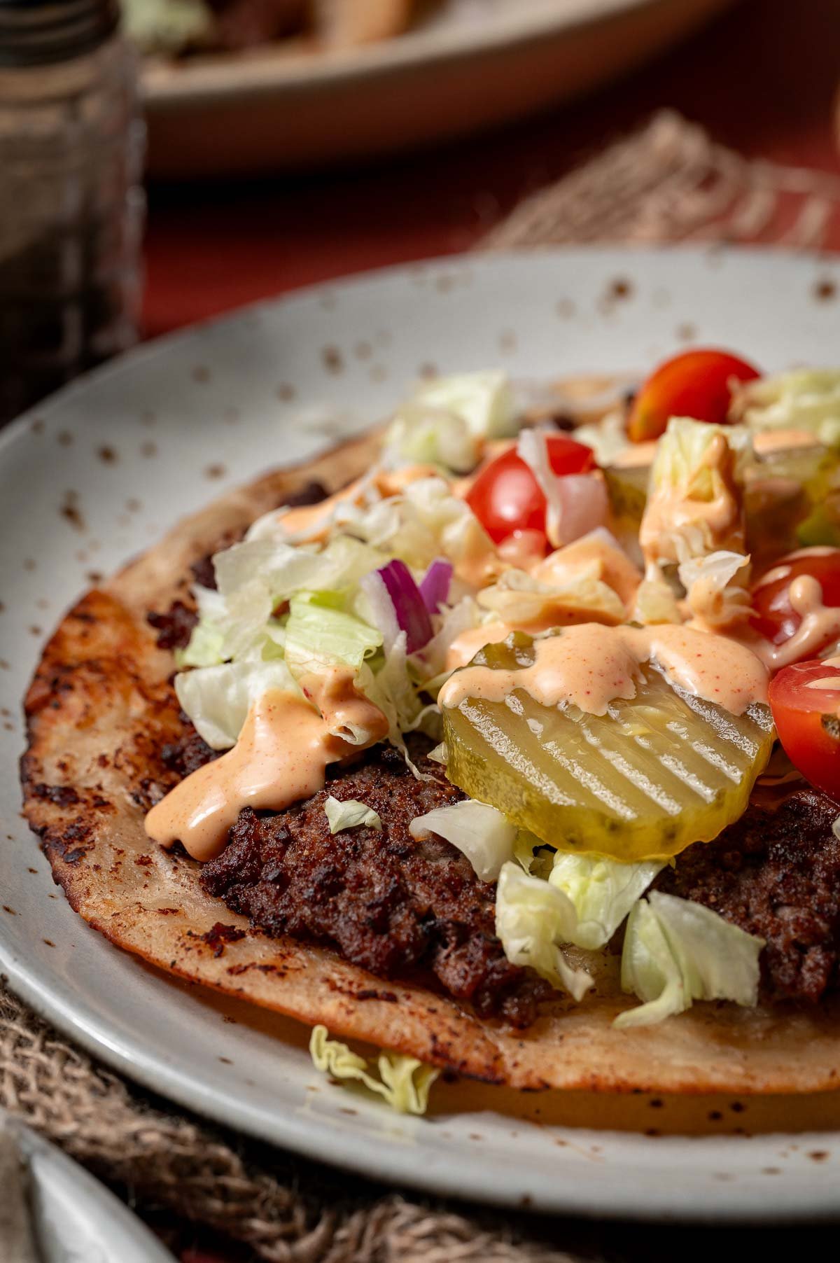 A plate with a smash burger taco topped with ground beef, sliced pickles, shredded lettuce, chopped tomato, red onion, and a drizzle of special sauce.