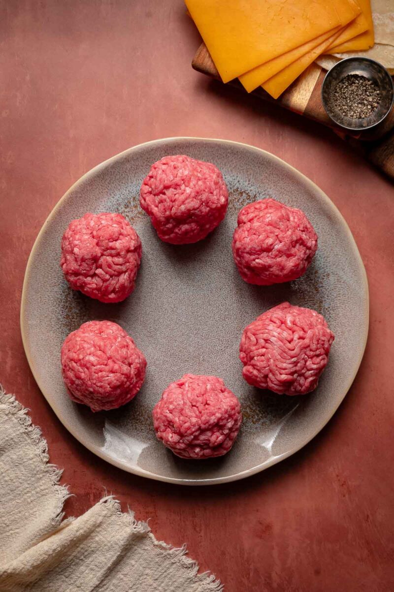 Six raw ground beef balls are arranged in a circle on a gray plate; slices of cheddar cheese and a small bowl of pepper are nearby on a wooden board.