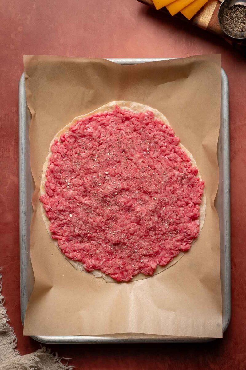 Raw ground beef spread on a tortilla, sprinkled with salt and pepper, placed on parchment paper on a baking sheet.