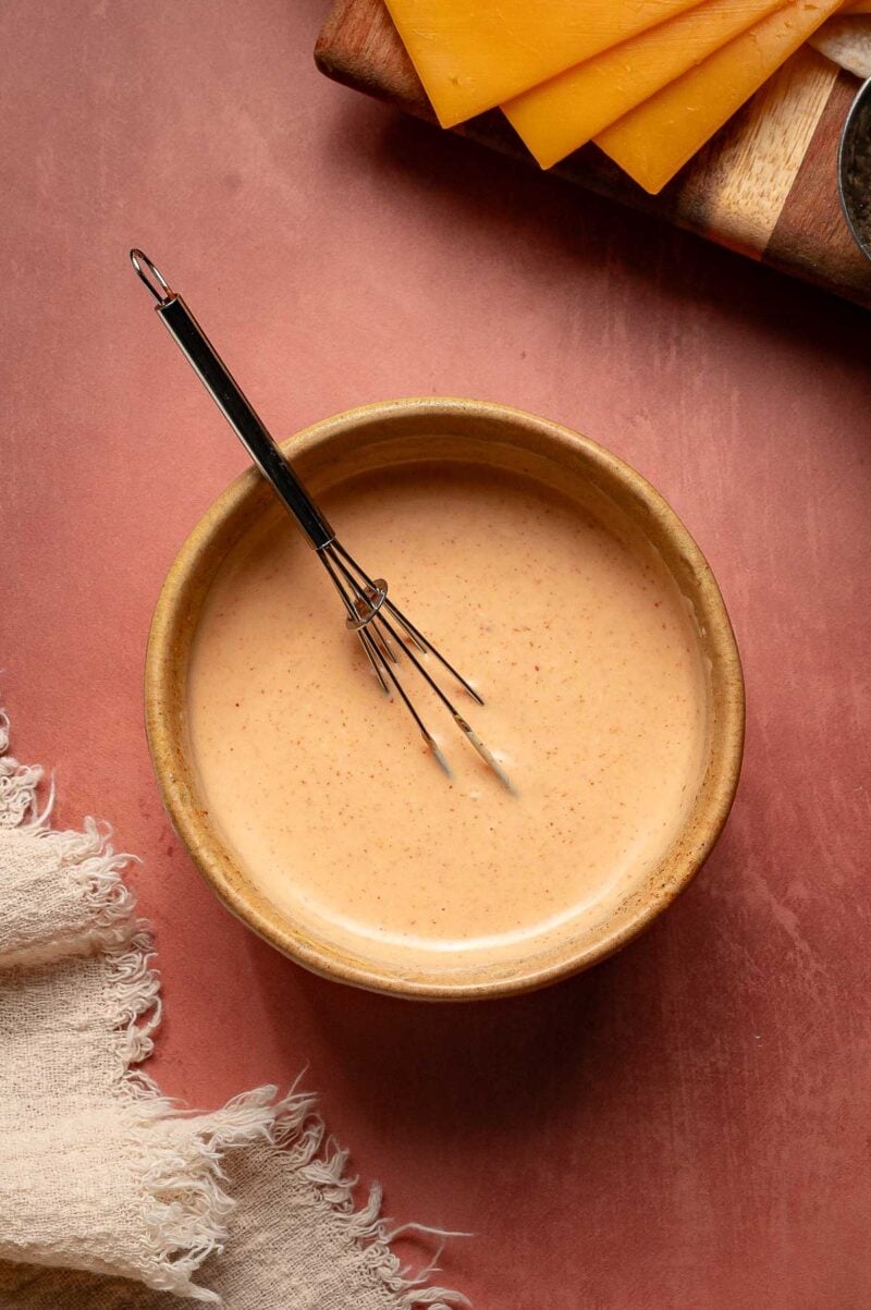 A small whisk rests in a bowl of creamy special sauce on a pink surface, with a beige cloth and sliced cheddar cheese nearby.