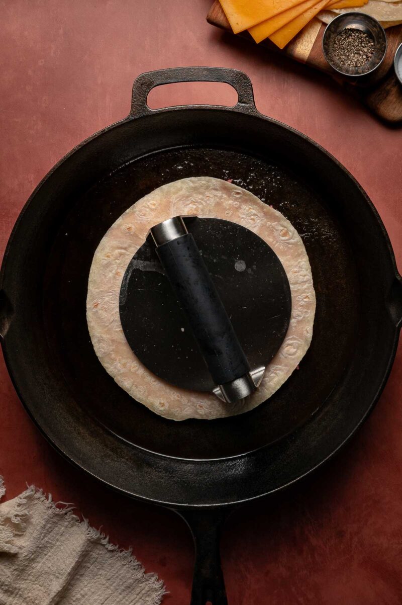 A smash burger taco is being pressed flat in a cast iron skillet with a metal tortilla press; cheese slices and seasonings are on a wooden board nearby.