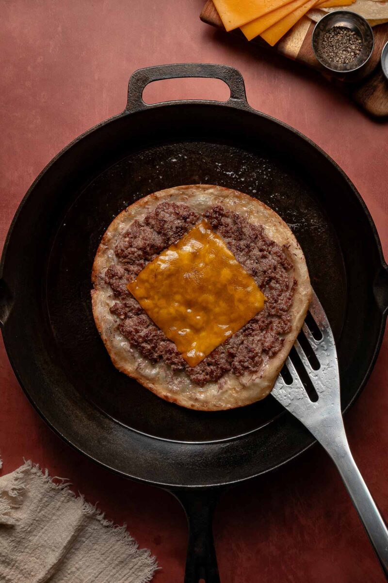 A cooked smash burger topped with ground beef and a slice of melted cheddar cheese sits in a cast-iron skillet with a metal spatula underneath.