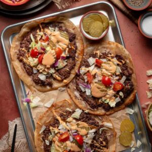 Three smash burger tacos topped with ground beef, shredded lettuce, chopped tomatoes, diced onions, pickles, and special sauce on a metal tray, with bowls of toppings and sauce nearby.