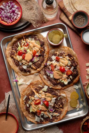 Three smash burger tacos topped with ground beef, shredded lettuce, chopped tomatoes, diced onions, pickles, and special sauce on a metal tray, with bowls of toppings and sauce nearby.