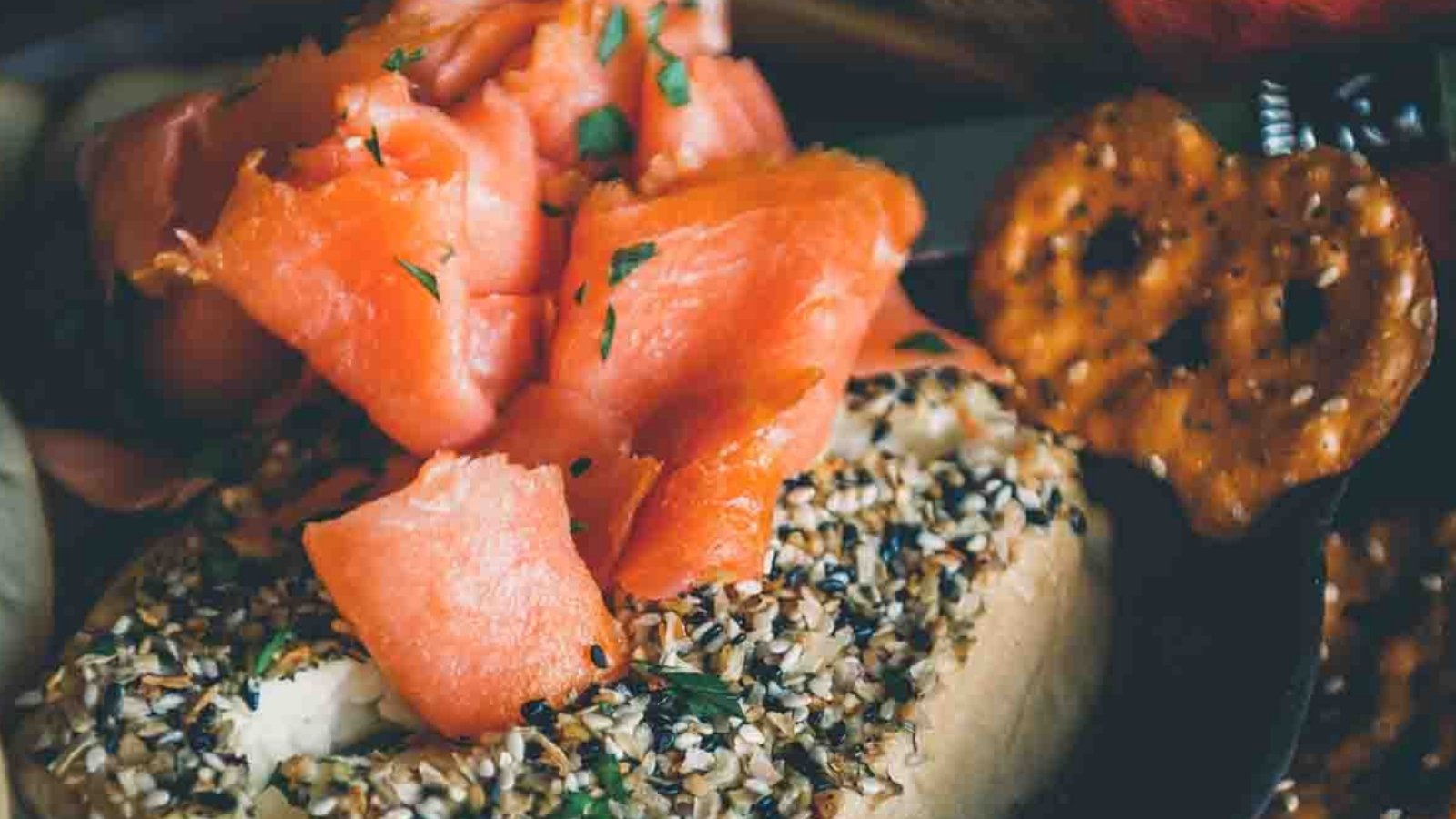 An everything bagel with cream cheese topped with slices of smoked salmon, served next to a seasoned pretzel crisp.