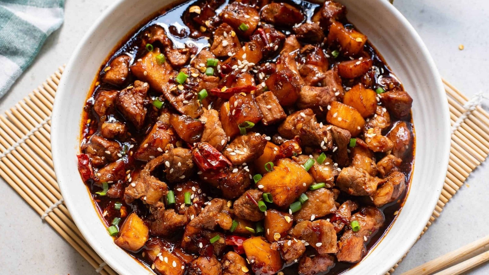 A bowl of cooked pork with pineapple chunks, red peppers, and green onions in a dark sauce, topped with sesame seeds.