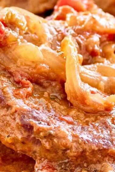 Close-up of a cooked beef steak topped with saut&eacute;ed onions and tomatoes in a rich, saucy mixture.