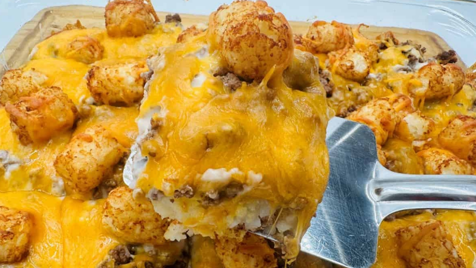 A close-up of a baked casserole topped with tater tots, ground beef, and melted cheddar cheese, with a serving spatula lifting a portion.
