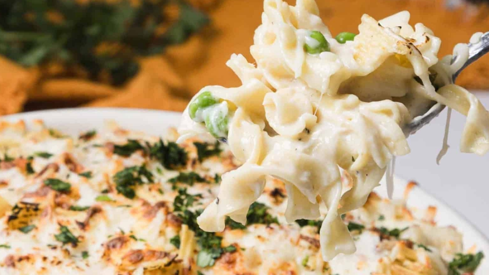 A fork lifts creamy pasta bake with peas and cheese from a casserole dish, garnished with herbs.