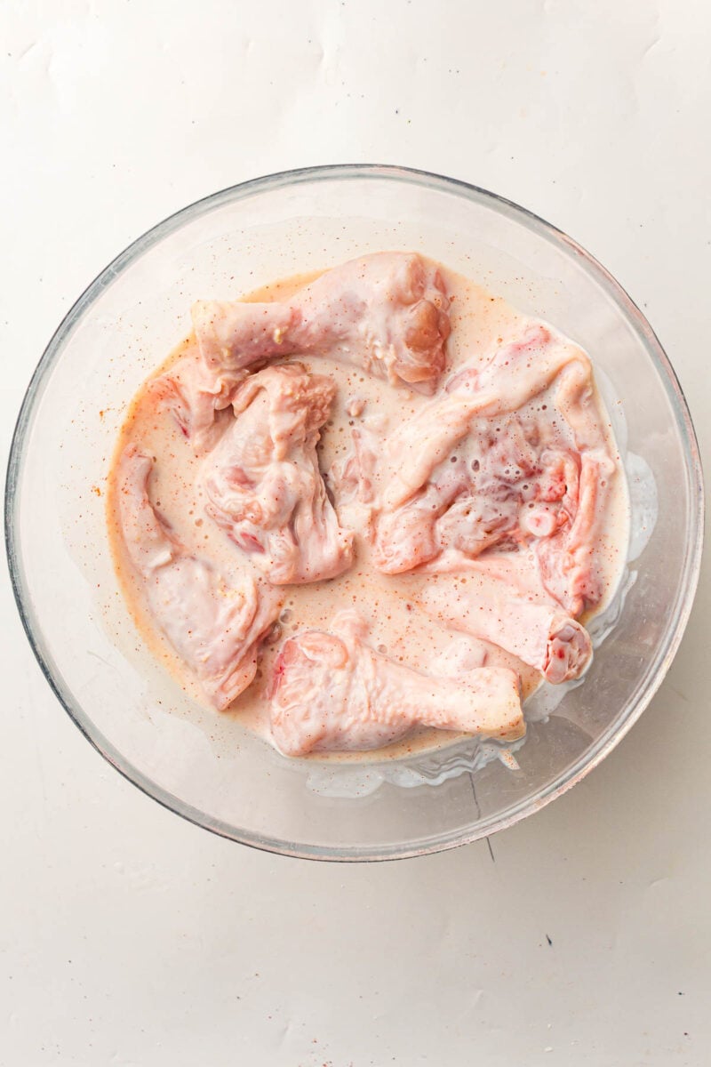 Raw chicken pieces marinating in a buttermilk with spices in a clear glass bowl on a light surface.