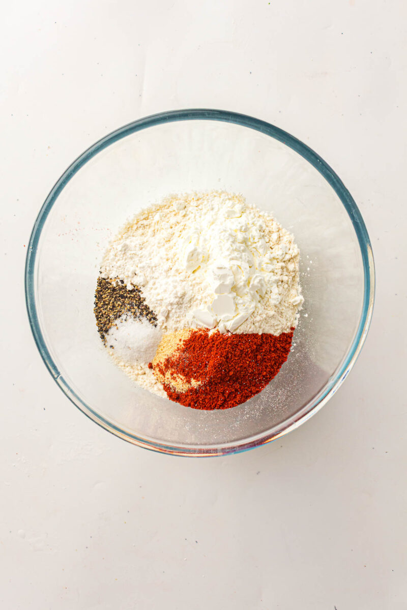 Clear glass bowl containing flour, black pepper, salt, paprika, and cornstarch, arranged in separate piles on a white surface.