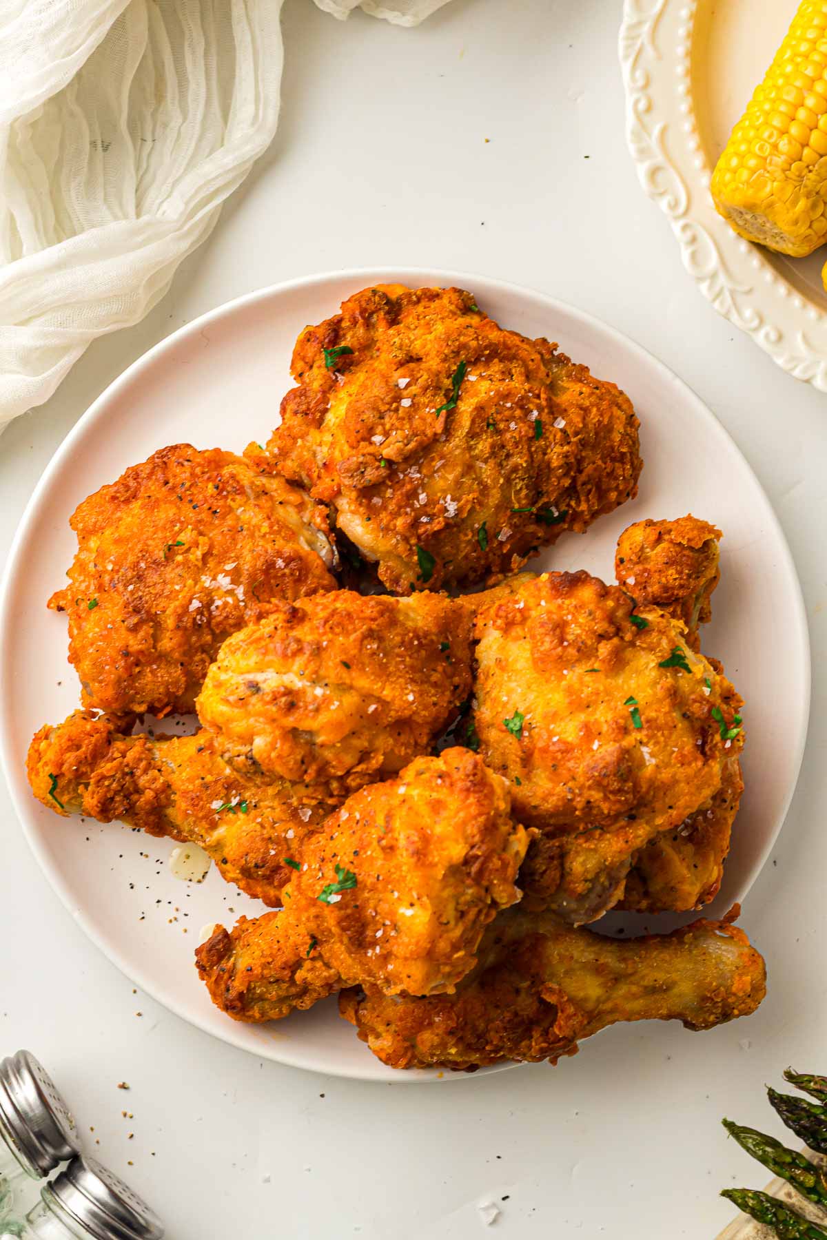 A white plate with several pieces of crispy air fryer fried chicken garnished with herbs, next to a yellow corn cob on a separate plate.