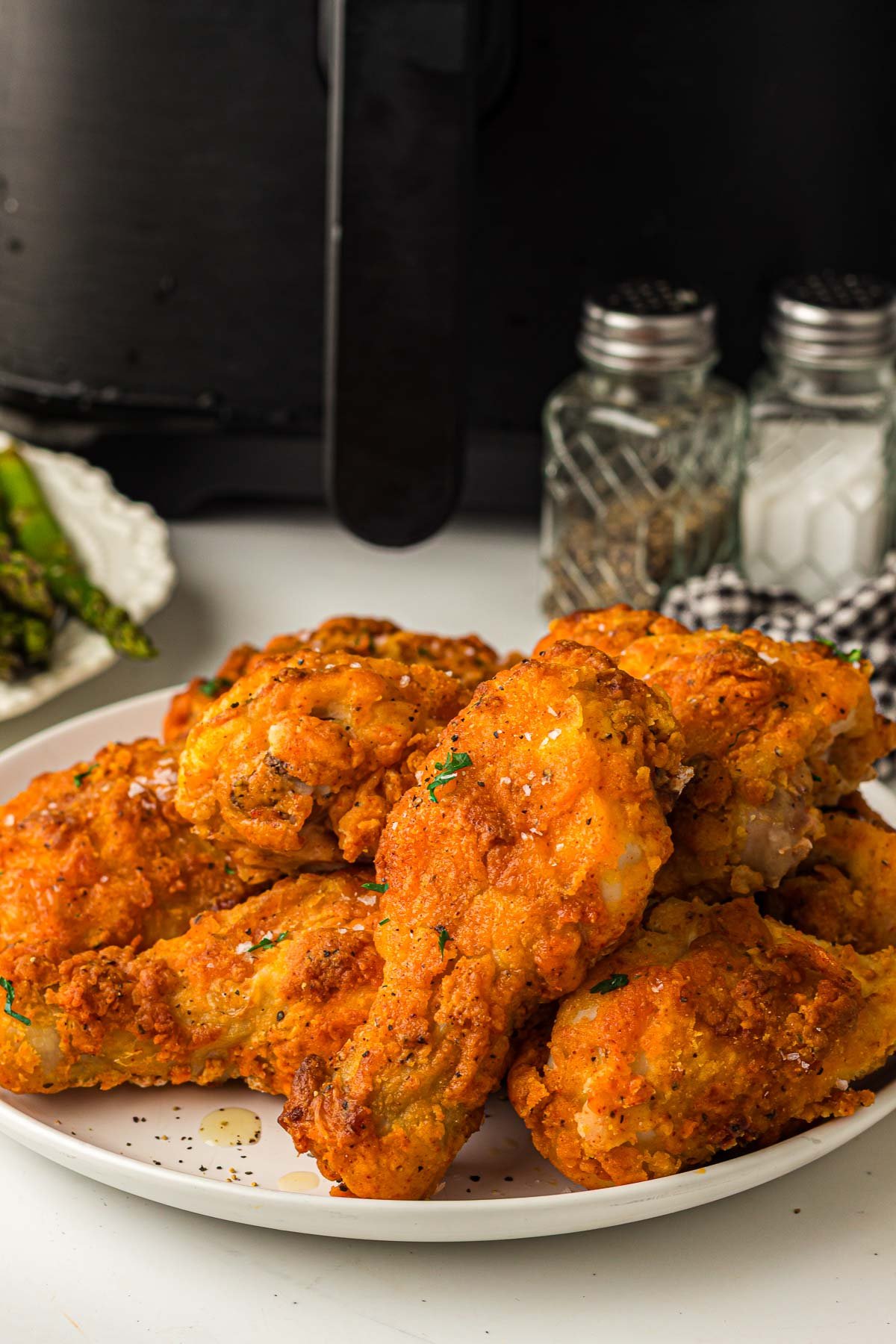 A plate of crispy fried chicken drumsticks is displayed in front of an air fryer, with salt and pepper shakers in the background.