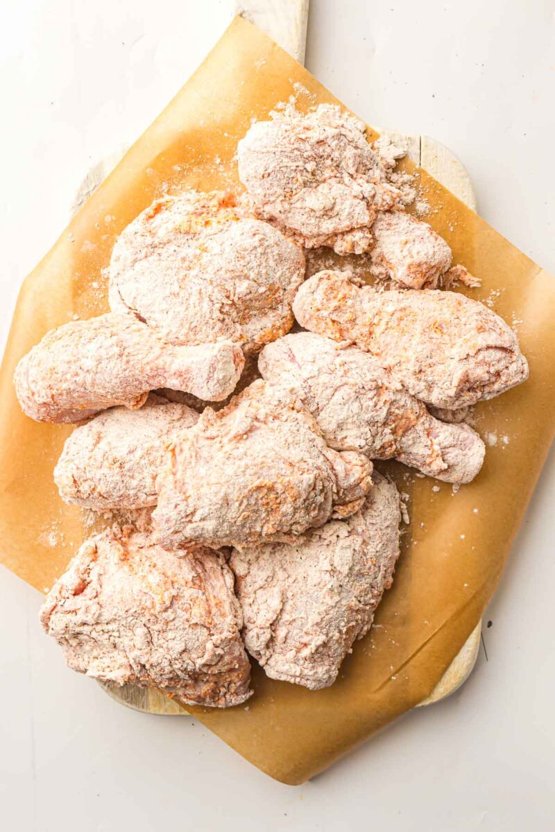 Raw chicken pieces coated in flour and seasoning are arranged on a sheet of brown parchment paper on a wooden board.
