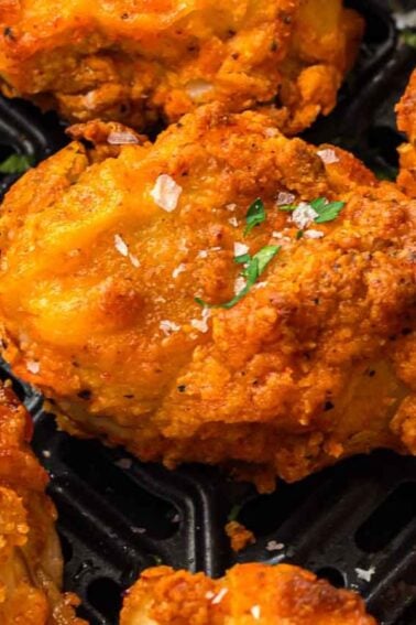 Close-up of crispy, golden-brown fried chicken pieces garnished with chopped herbs and coarse salt on a dark tray.