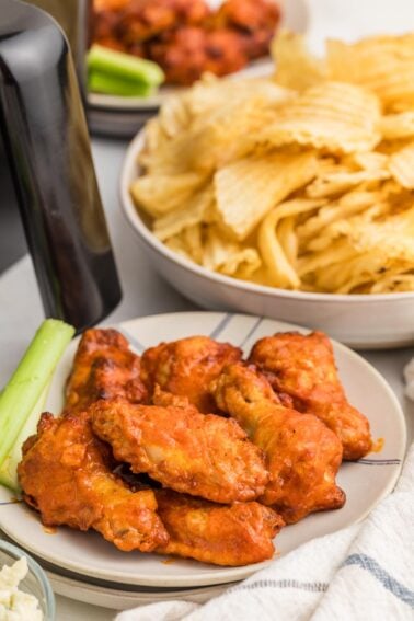 Plate of buffalo chicken wings with a celery stick, next to a bowl of ridged potato chips and another plate of food in the background.