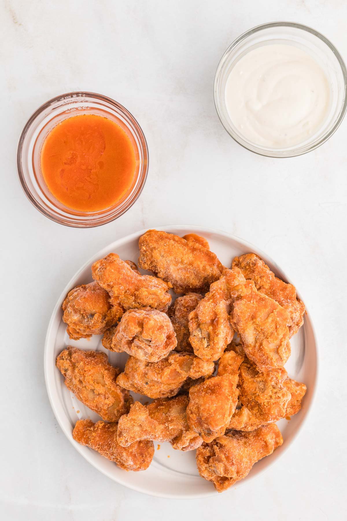 A plate of breaded chicken wings is next to a bowl of buffalo sauce and a bowl of ranch dressing on a white surface.
