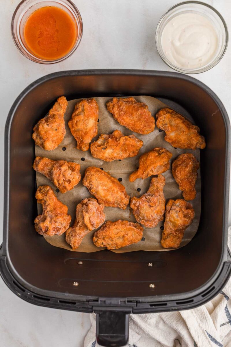 Frozen chicken wings arranged in a single layer inside an air fryer basket lined with parchment paper, with bowls of sauce and dip nearby.