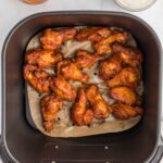 Chicken wings in an air fryer basket on parchment paper, with bowls of buffalo sauce and ranch dressing nearby.