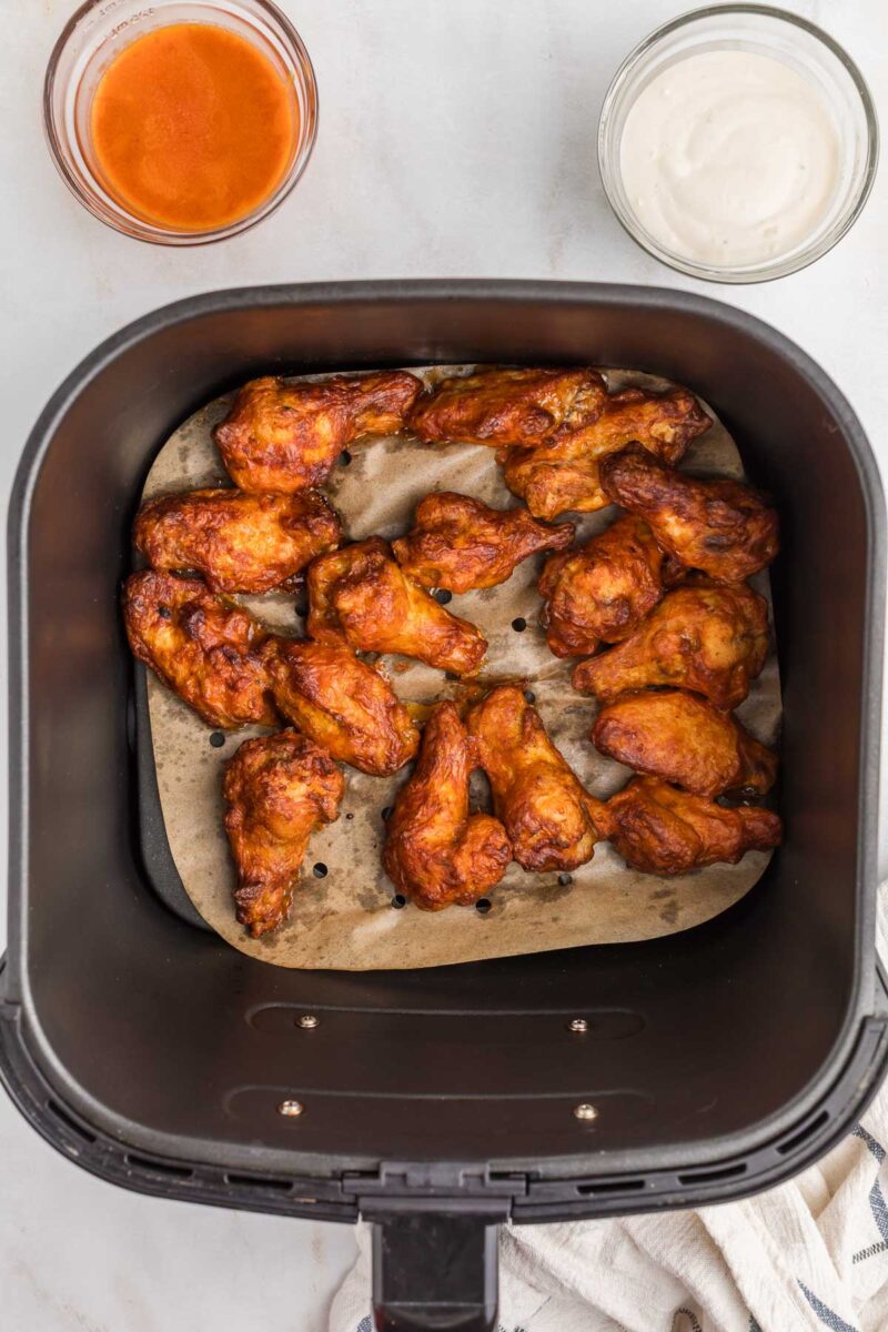 Chicken wings in an air fryer basket on parchment paper, with bowls of buffalo sauce and ranch dressing nearby.