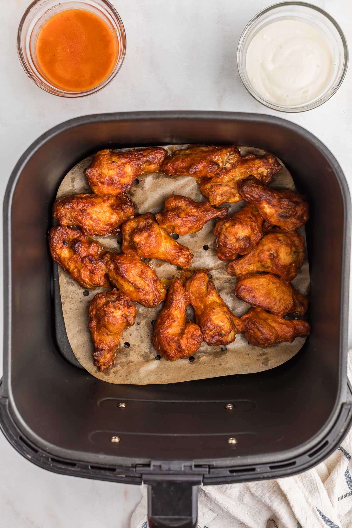 Chicken wings in an air fryer basket on parchment paper, with bowls of buffalo sauce and ranch dressing nearby.