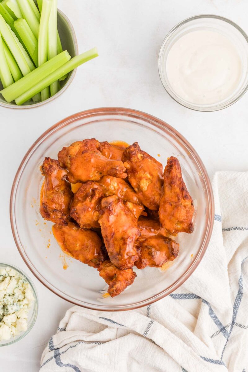 A glass bowl of buffalo chicken wings, a bowl of celery sticks, a bowl of blue cheese crumbles, and a bowl of ranch dressing on a white surface with a cloth napkin.