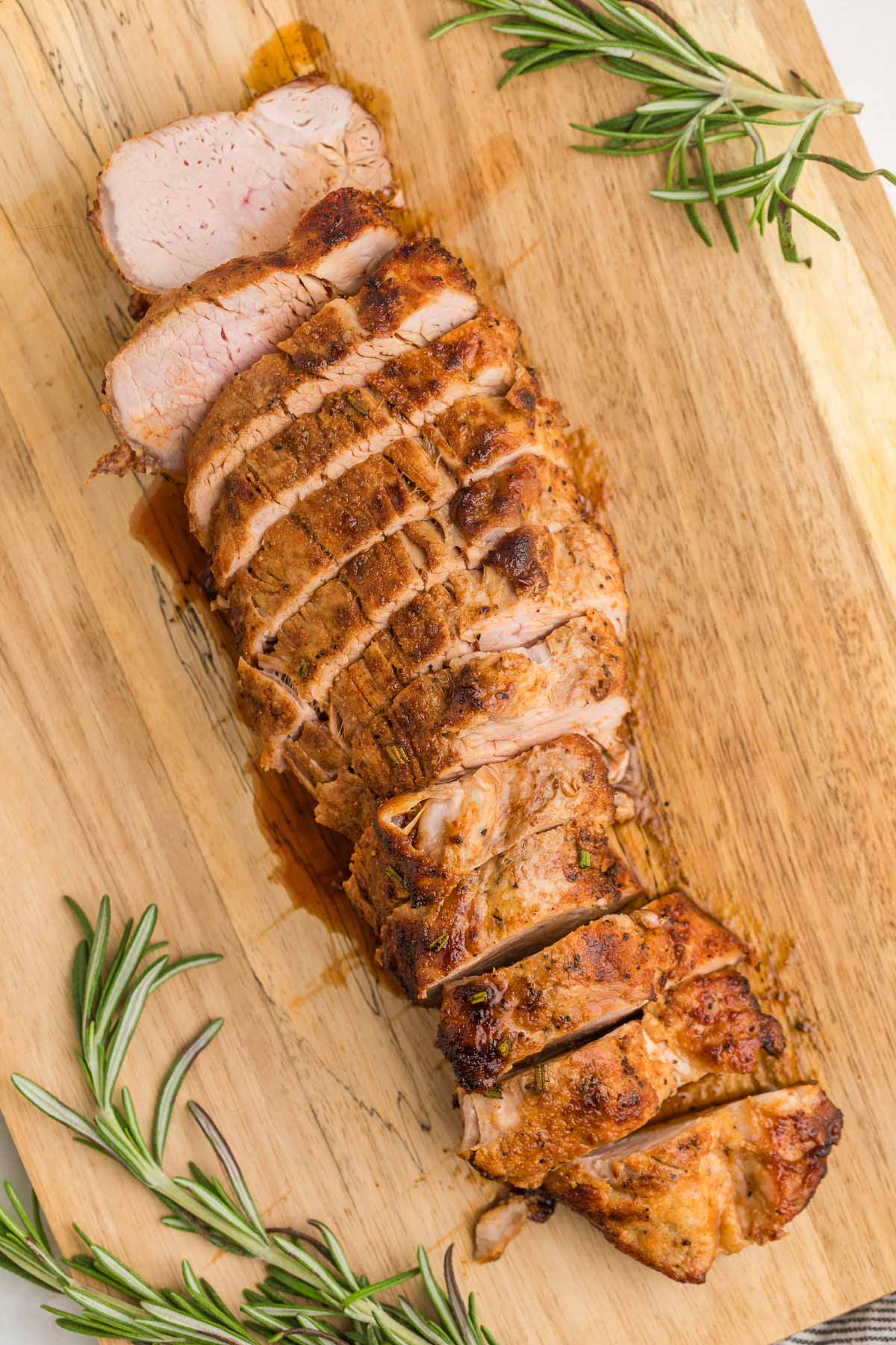 Sliced air fryer pork tenderloin on a wooden cutting board, garnished with fresh rosemary sprigs.