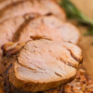 Close-up of several slices of cooked air fryer pork tenderloin, arranged on a wooden cutting board with sprigs of rosemary in the background.