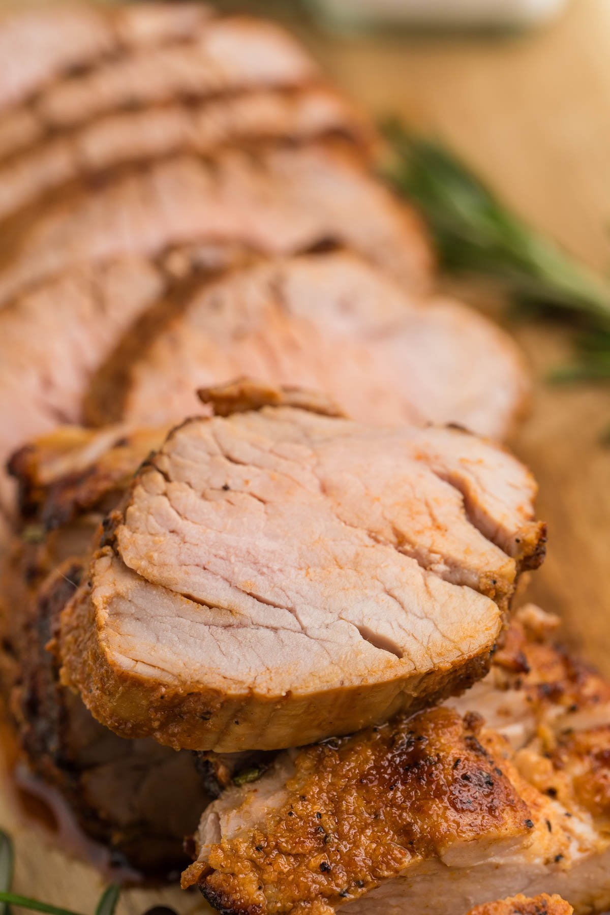 Close-up of several slices of cooked air fryer pork tenderloin, arranged on a wooden cutting board with sprigs of rosemary in the background.