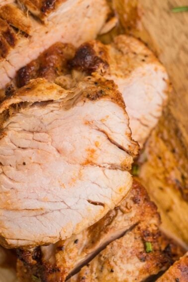 Sliced cooked pork tenderloin arranged on a wooden cutting board with sprigs of rosemary beside it.