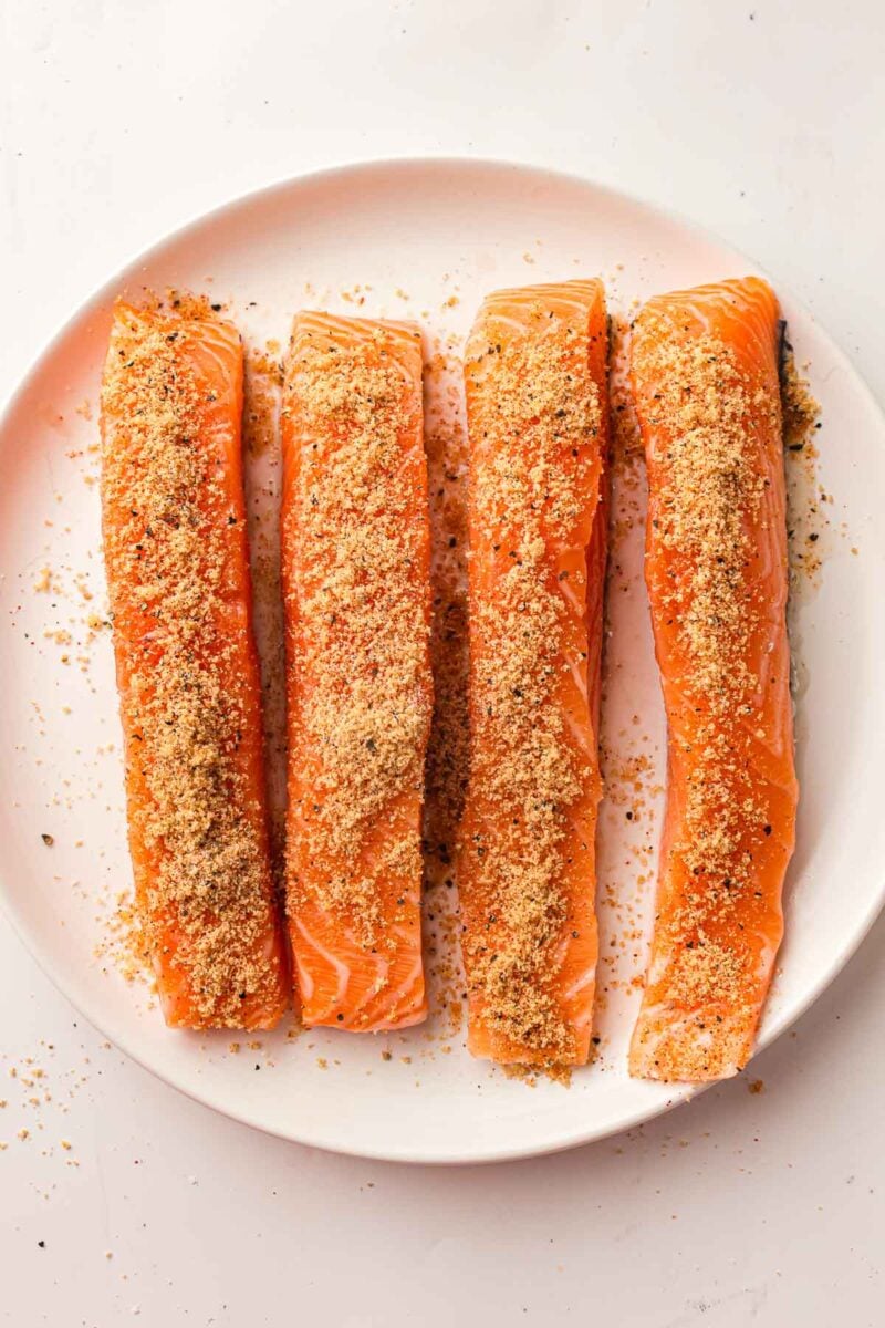 Four raw salmon fillets on a white plate, topped with a layer of seasoned breadcrumbs.