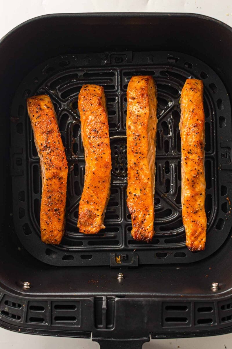 Four seasoned salmon fillets are arranged in a single layer inside an air fryer basket, cooked with a golden-brown finish.