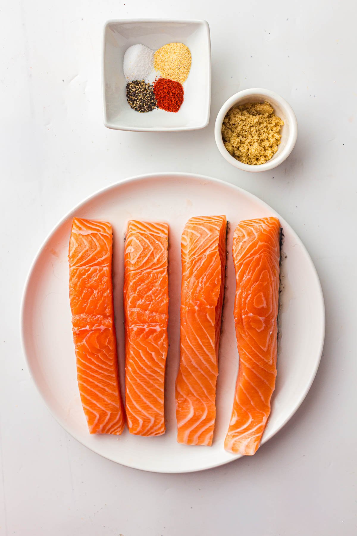 Four raw salmon fillets on a white plate, with a small bowl of brown sugar and a separate dish containing assorted spices on a white surface.