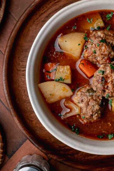 A bowl of meatball soup with potatoes, carrots, and herbs beside a drink with lemon slices on a wooden table.