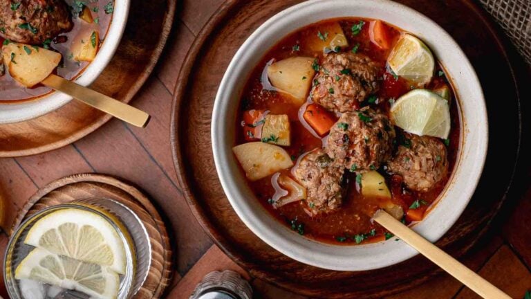 A bowl of meatball soup with potatoes, carrots, and herbs beside a drink with lemon slices on a wooden table.
