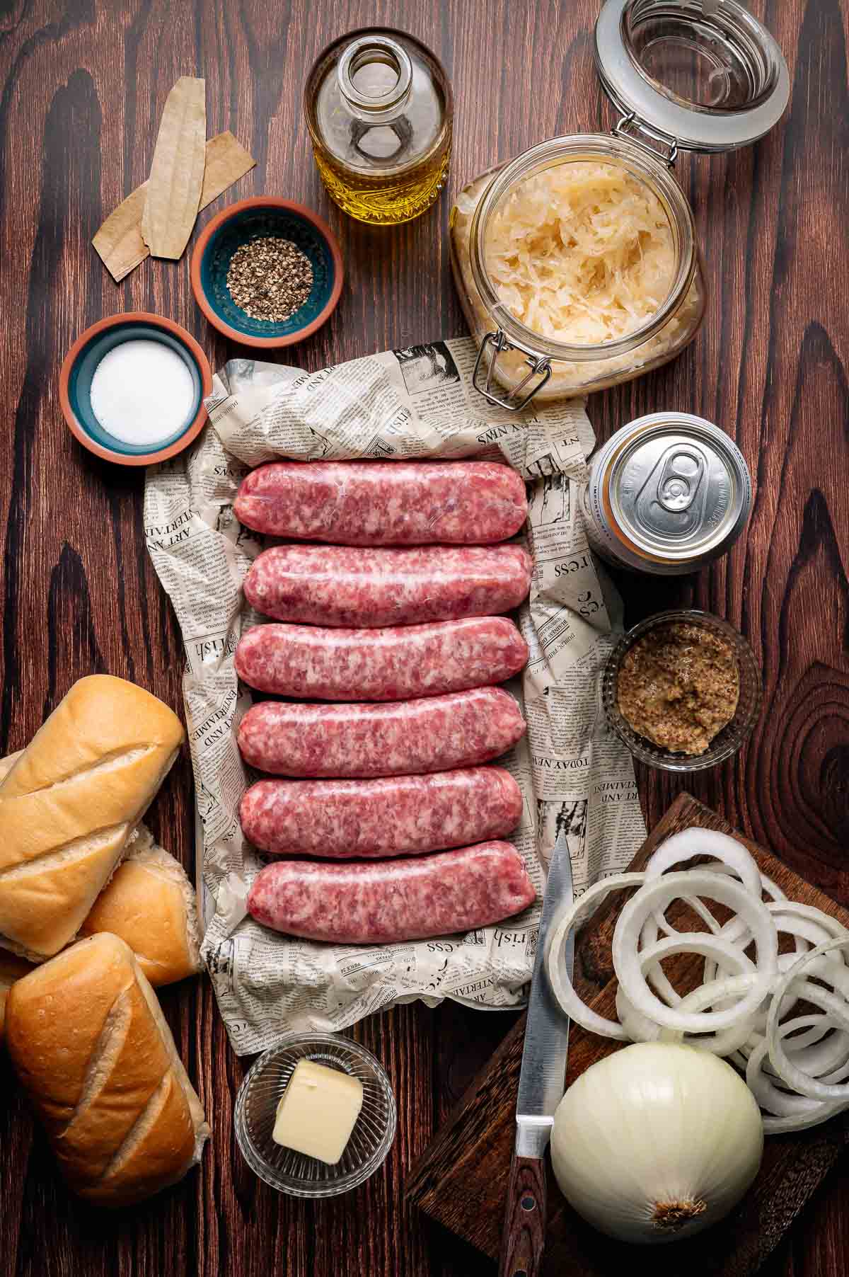 Uncooked sausages on paper, surrounded by buns, sliced onions, sauerkraut, butter, seasonings, oil, brown sugar, and a can of beer on a wooden table.