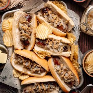 Tray of beer brats in buns topped with onions and grainy mustard, surrounded by ridged potato chips on a rustic table setting.
